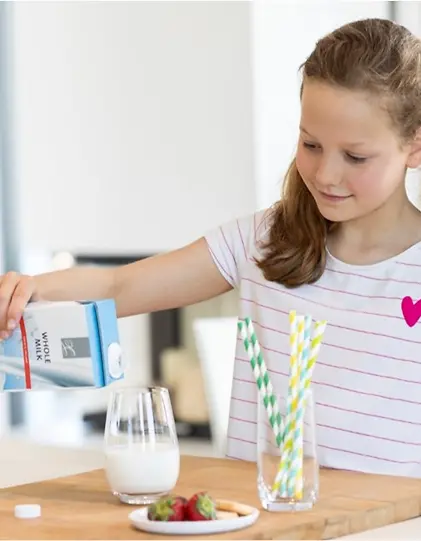 A girl pouring milk into a cup, with a glass of paper straws beside it.