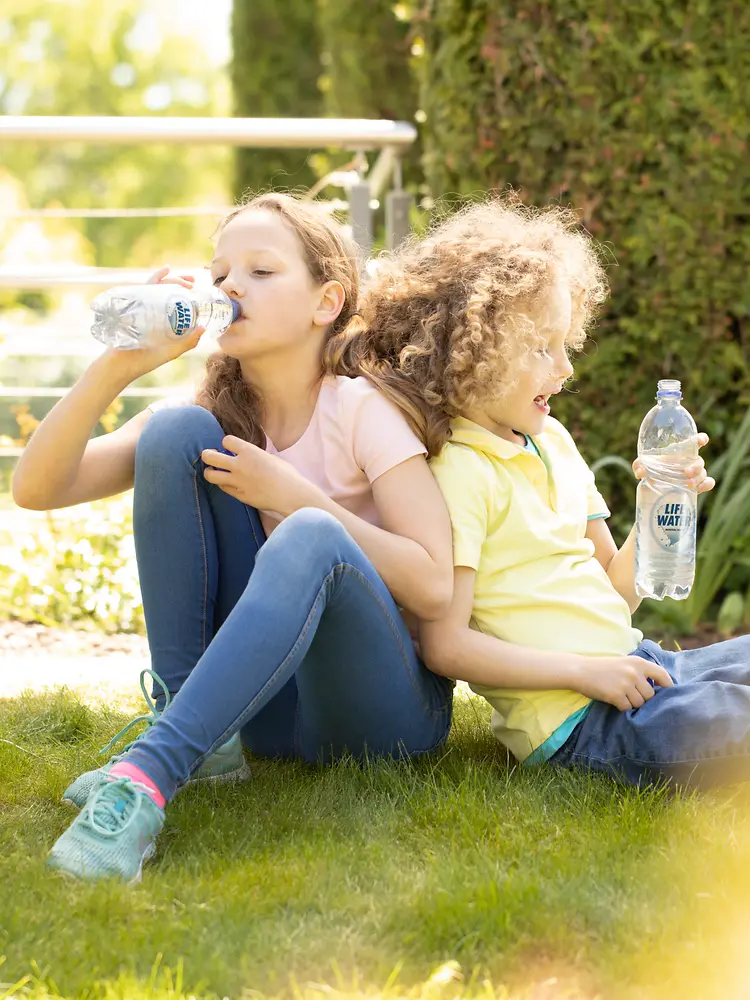 Two children drinking bottled water on the grass.