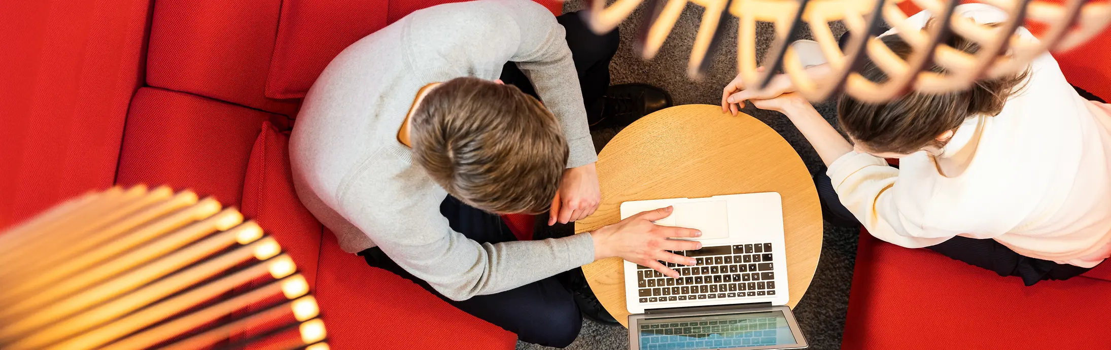 Dua orang duduk di sofa merah dan bekerja bersama di laptop